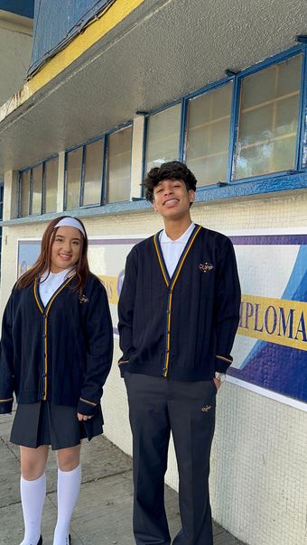 Dos personas con cárdigans y faldas/pantalones a juego sonríen cerca de un edificio con el texto “Diploma”.