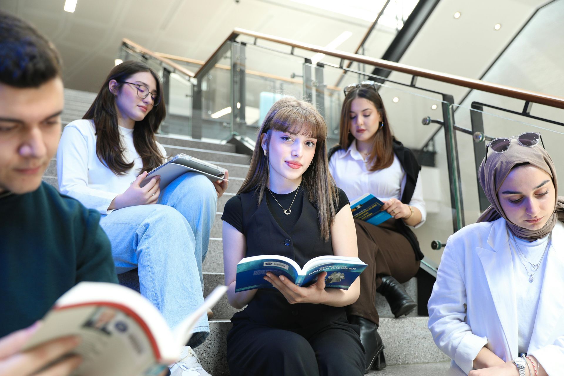 Estudiantes estudiando juntos en las escaleras, sosteniendo libros y una tableta en un espacio bien iluminado.