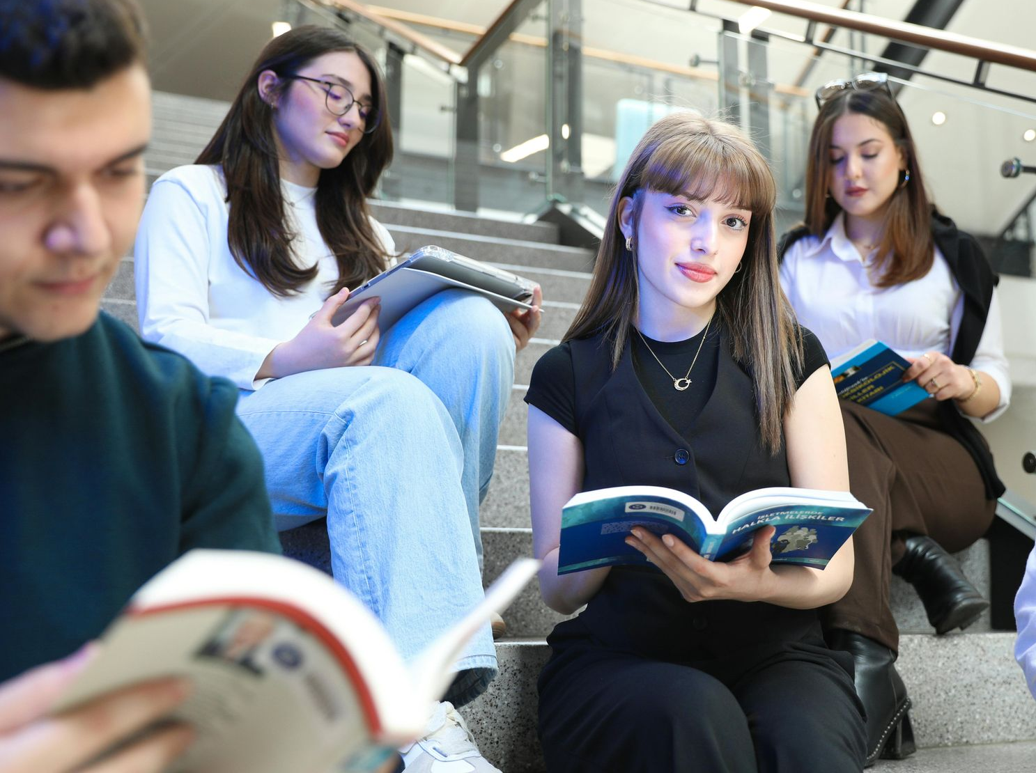 Estudiantes leyendo libros, sentados en las escaleras de un edificio. Uno mira a la cámara.