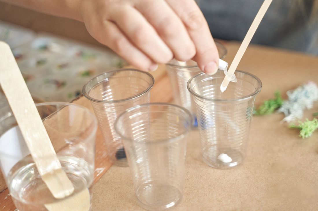 A person is mixing liquid in plastic cups with wooden sticks.