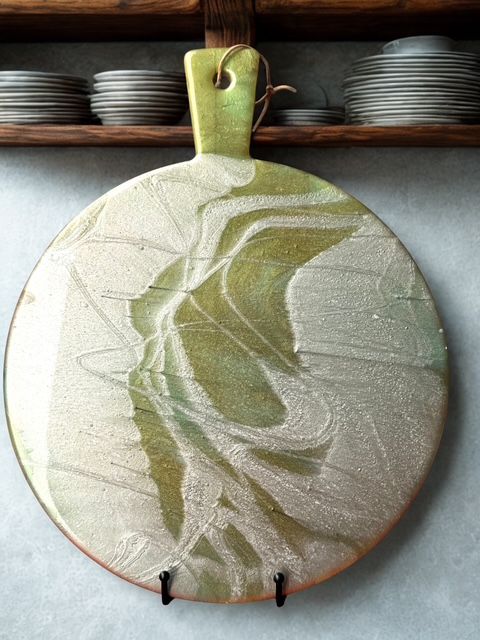 A round cutting board with a green handle is hanging on a shelf.