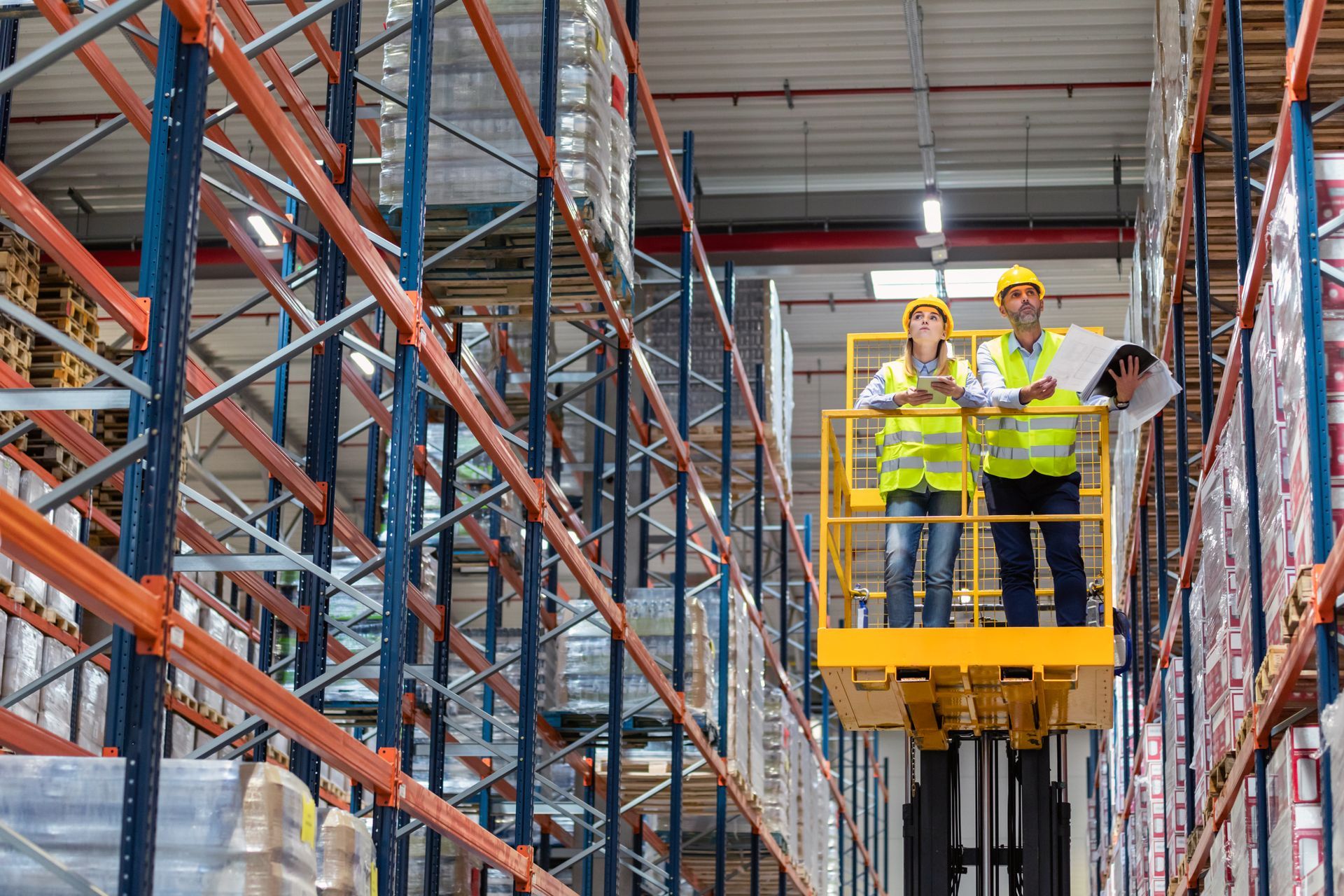 A man and a woman are standing on a forklift in a warehouse.