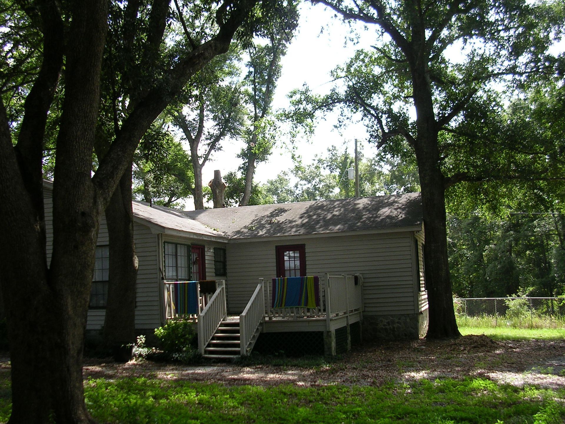 A white house with a deck nestled among large trees in a grassy, shaded yard.