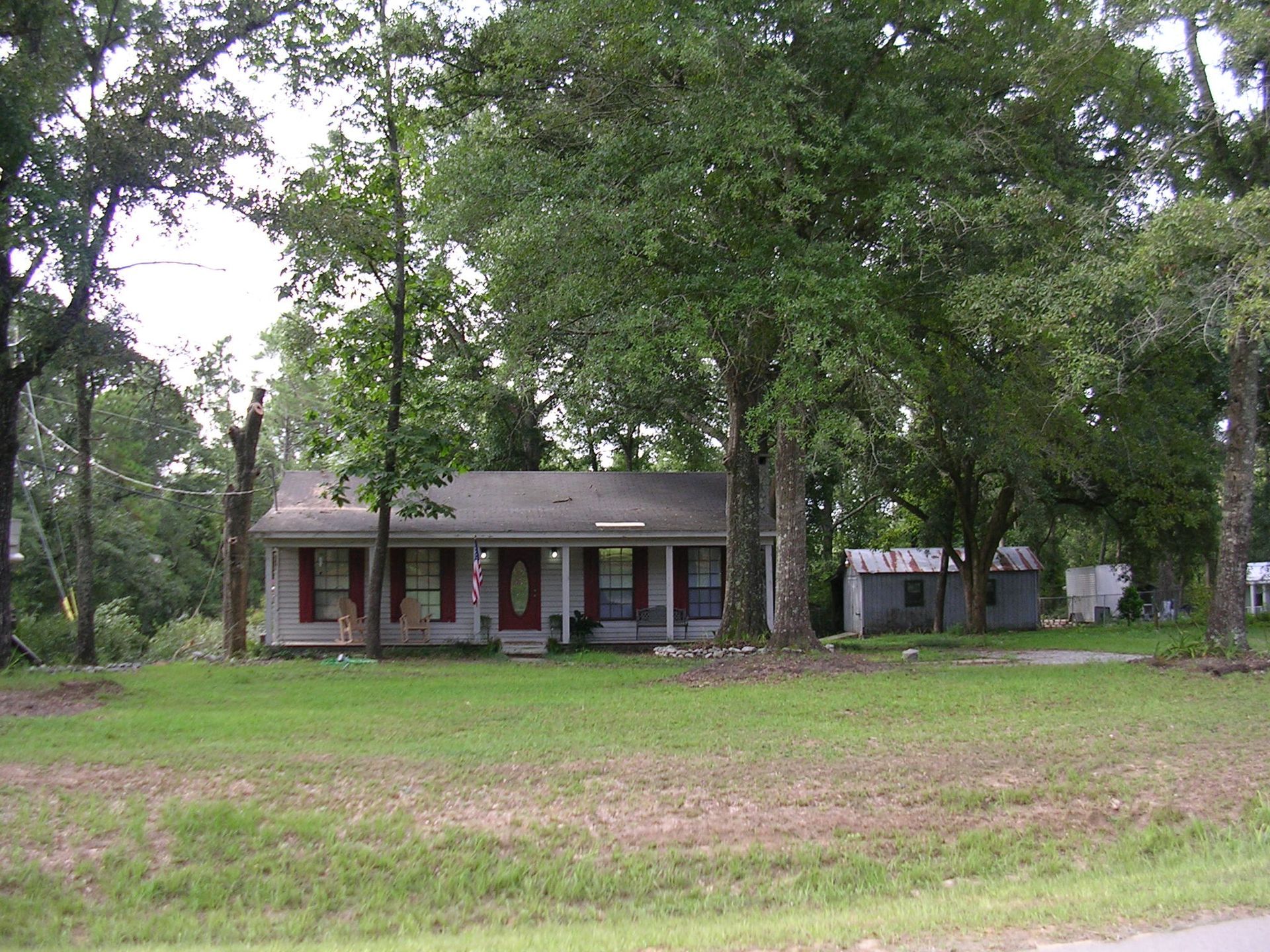 A single-story, light-colored house with dark red shutters sits behind a grassy yard, surrounded by numerous mature trees.