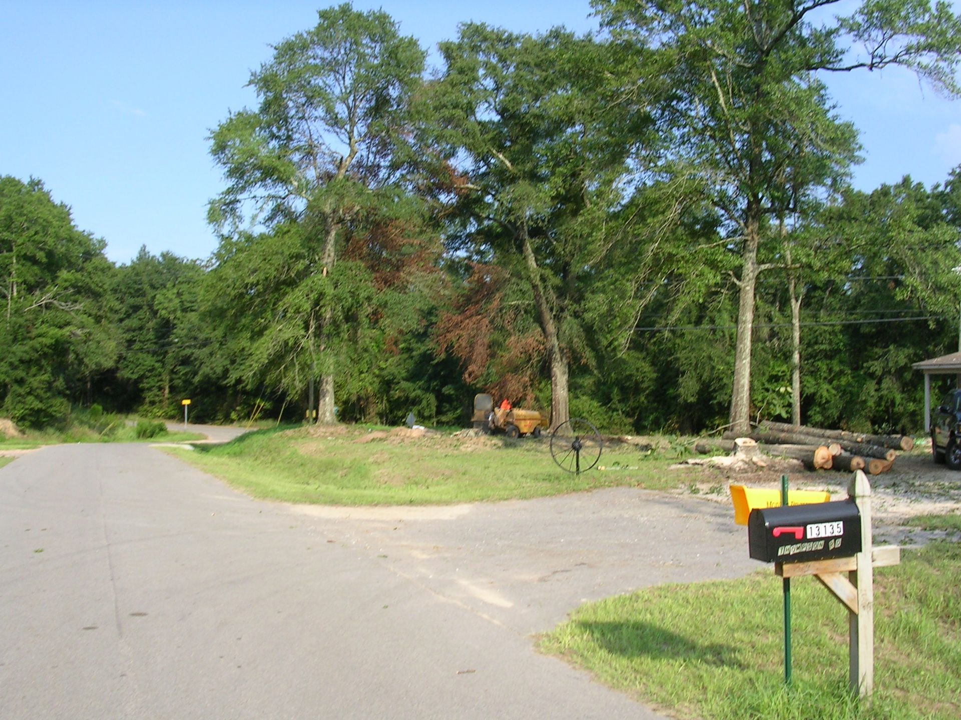 A black mailbox sits at the end of a gravel driveway near a road, with trees and a small tractor in the background.