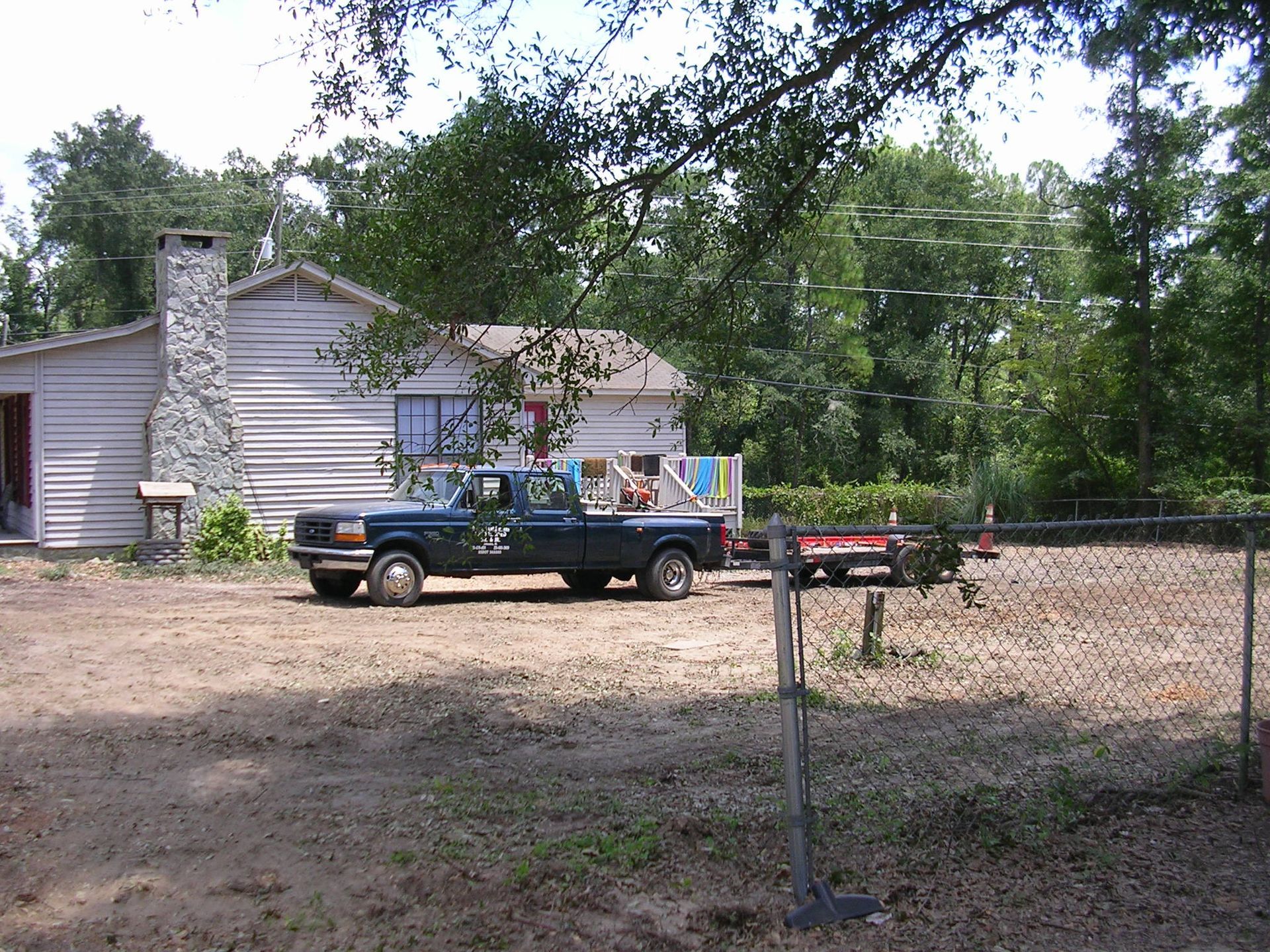 A dark pickup truck with a trailer parked in a dirt lot in front of a white house with a stone chimney.