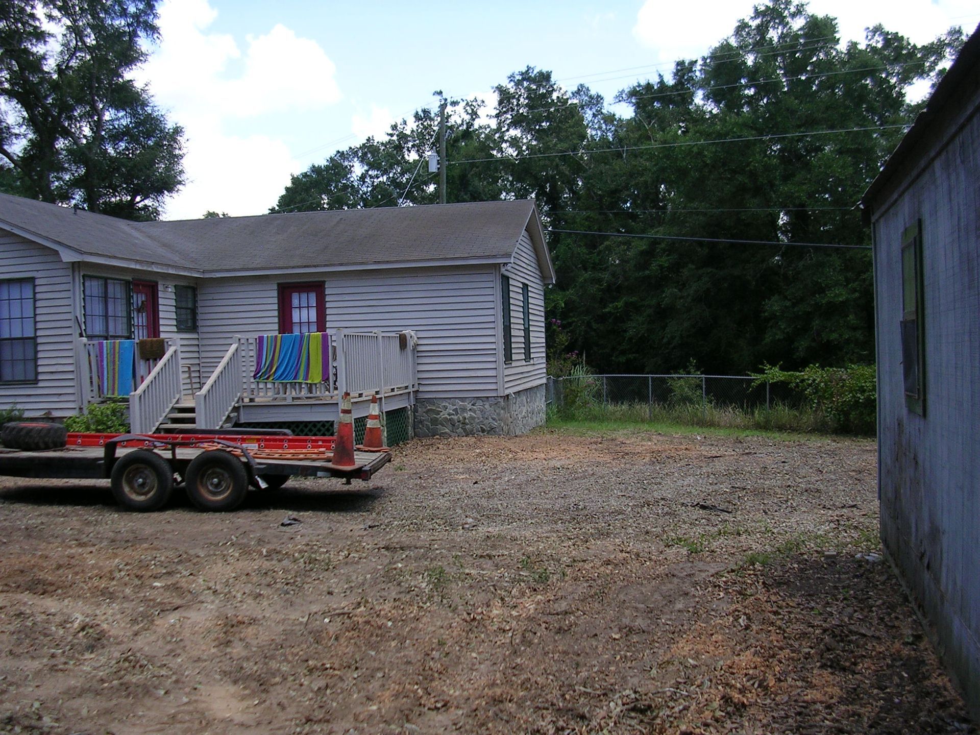 A light-colored house with a small porch and a utility trailer parked on a gravel driveway in a yard with trees.