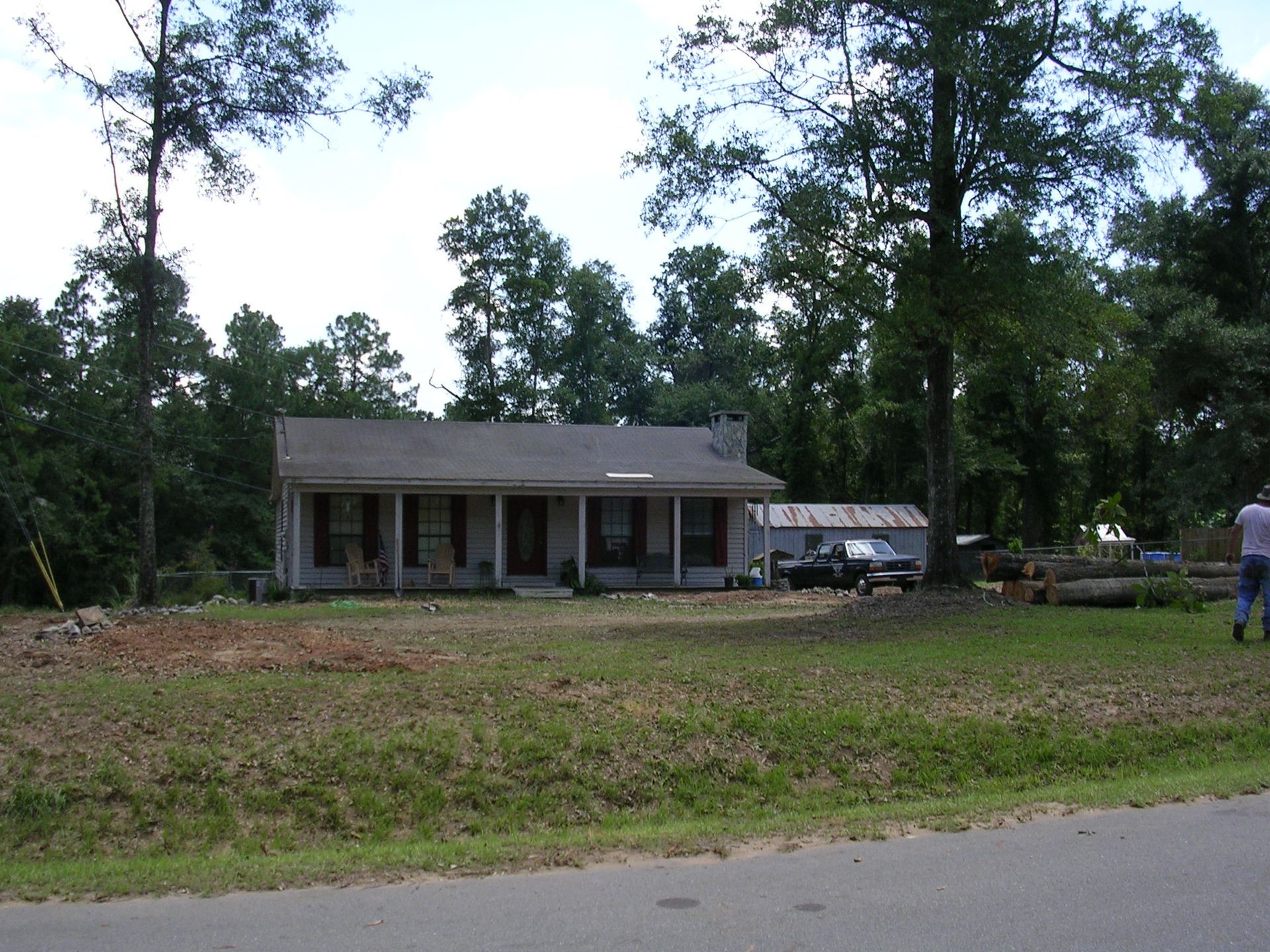 A light-colored house with a front porch, set back from a road behind a grassy lawn and surrounded by mature trees.