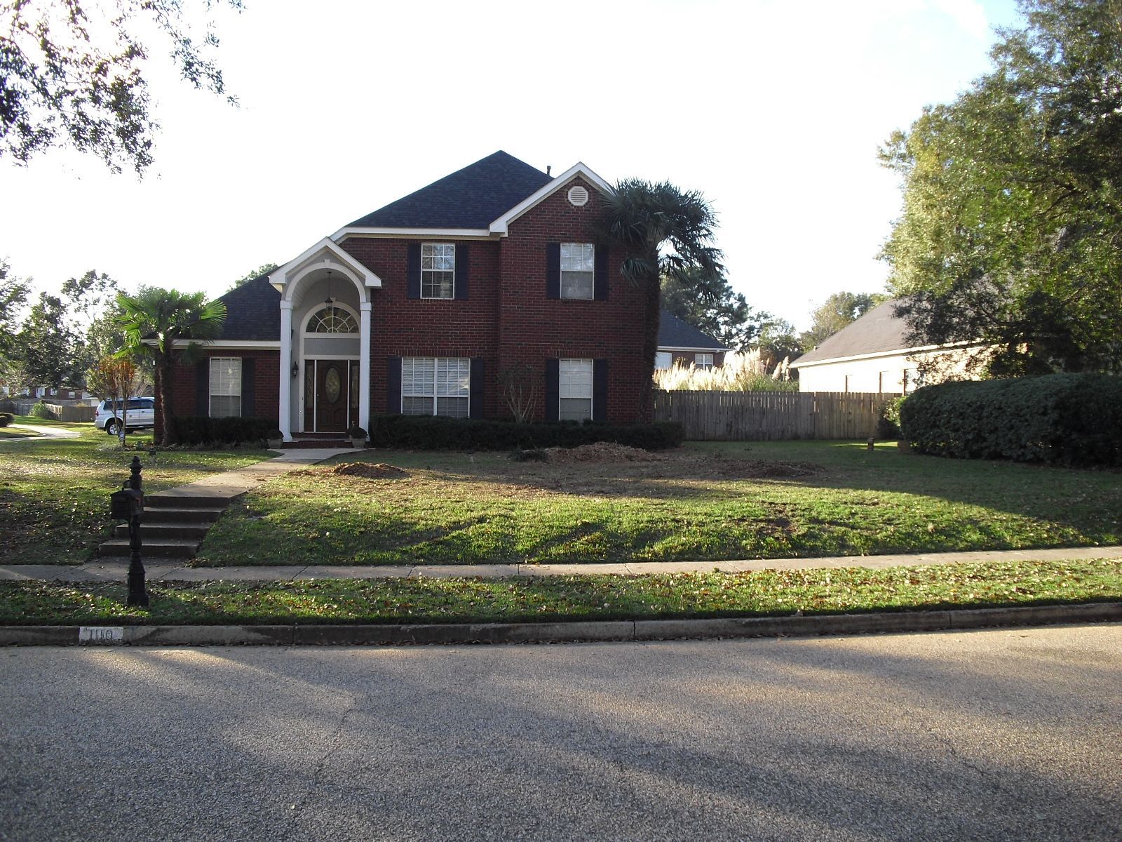 Brick two-story house with a black roof and arched entryway on a sunny day. A lawn and street are in front.