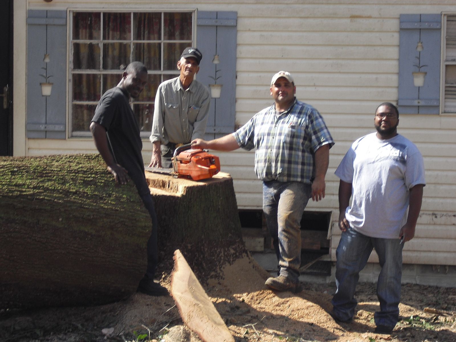 Four people stand in front of a house next to a large, freshly cut tree stump with a chainsaw resting on top.