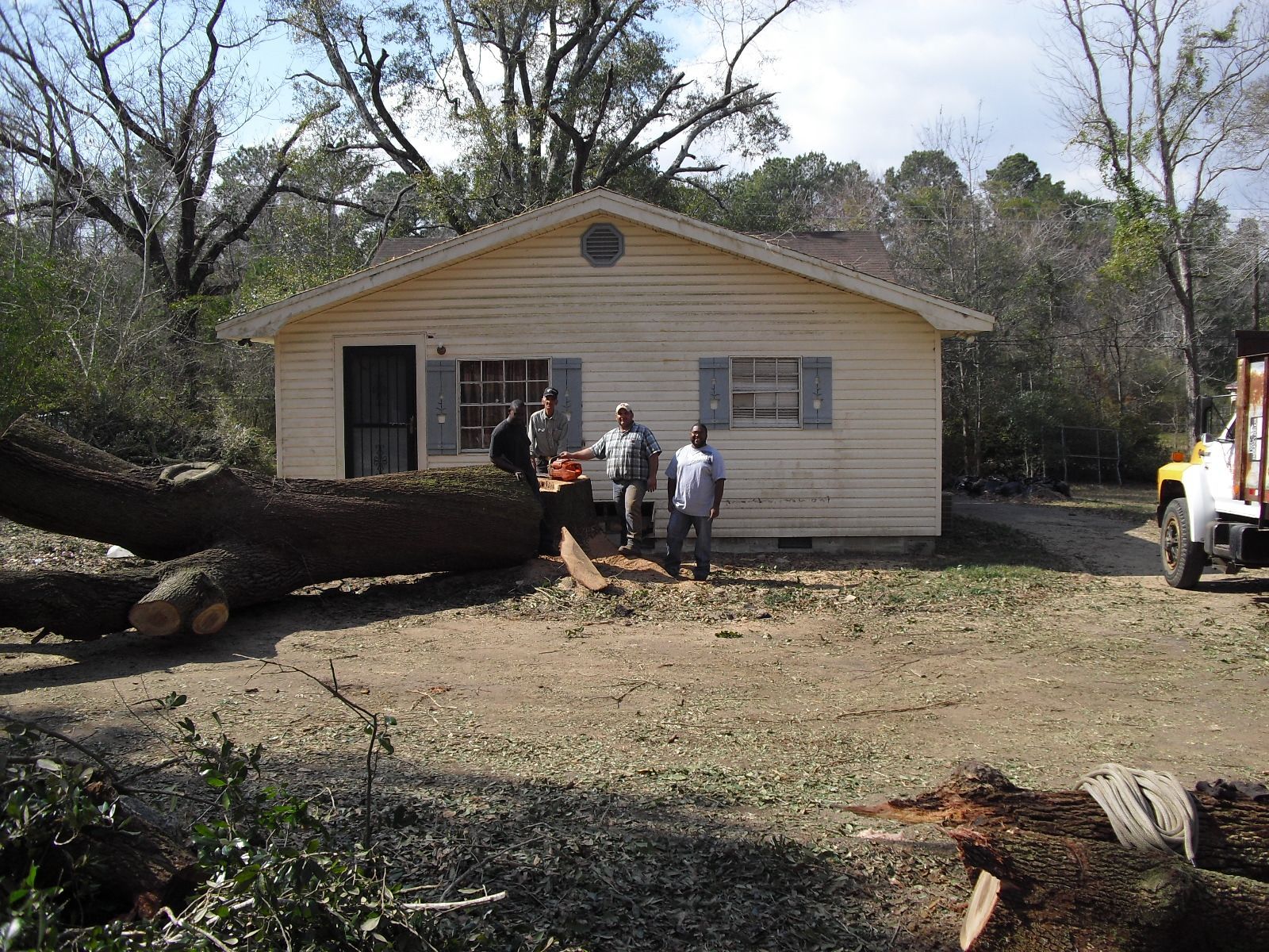 Three people stand in the yard of a light-colored house near a large fallen tree being cut with a chainsaw.