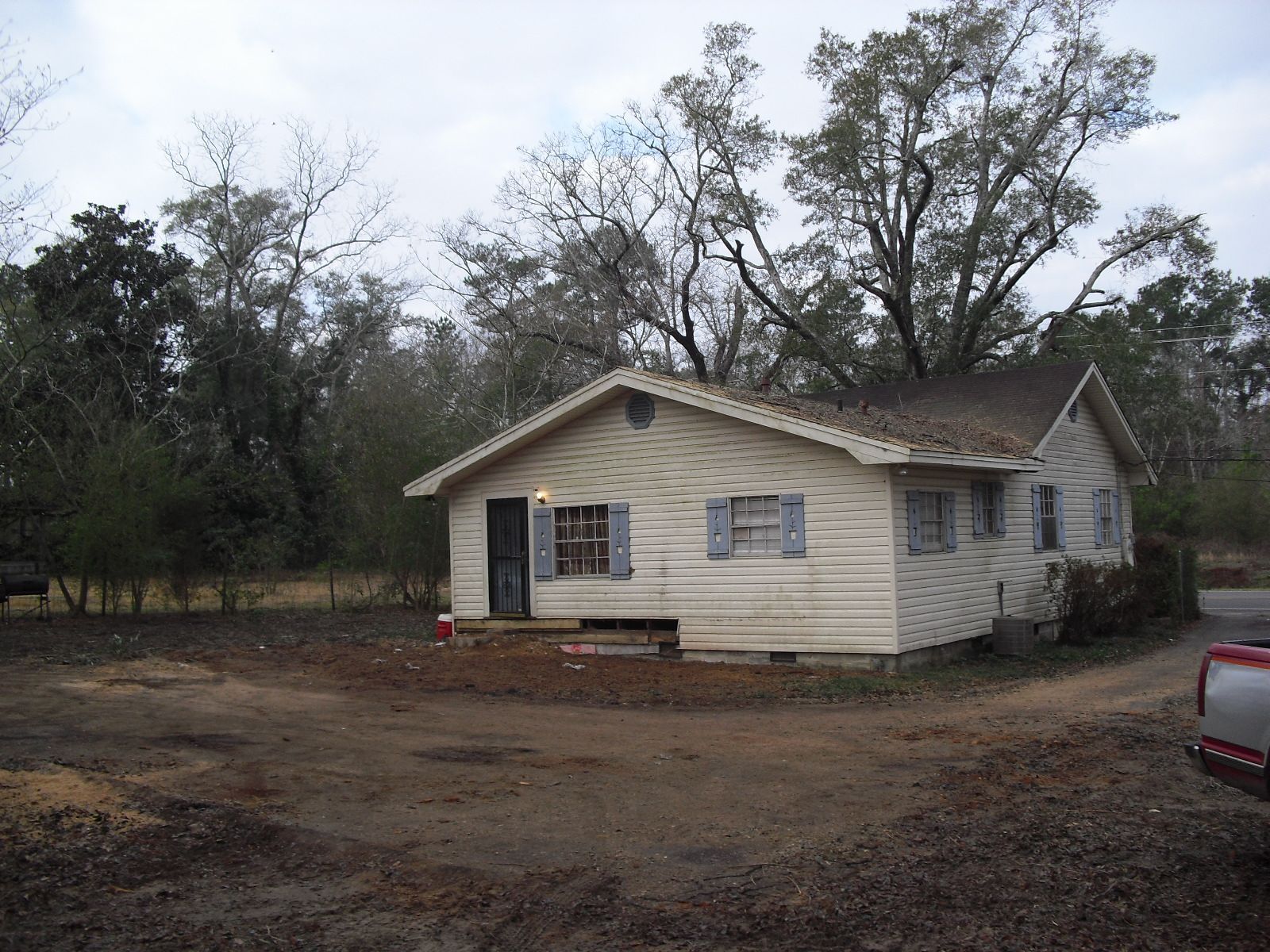 A single-story, light-colored wood siding house with blue shutters sits on a dirt lot surrounded by trees.