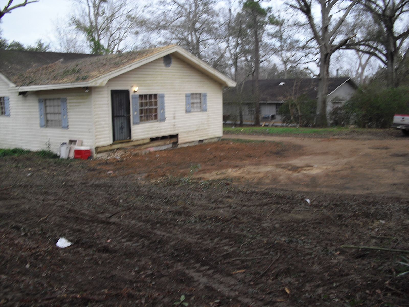 A white, single-story house with blue shutters sits on a dirt lot with a neighboring house visible in the background.