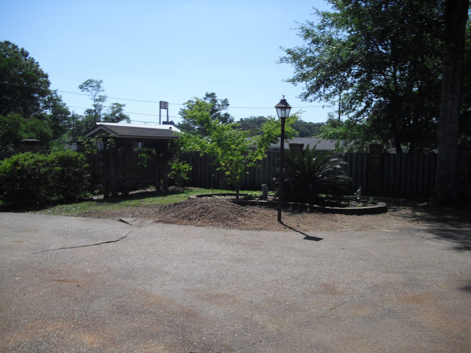 A gravel driveway leads to a landscaped yard with a small tree, a lamp post, and a wooden gazebo under a clear blue sky.
