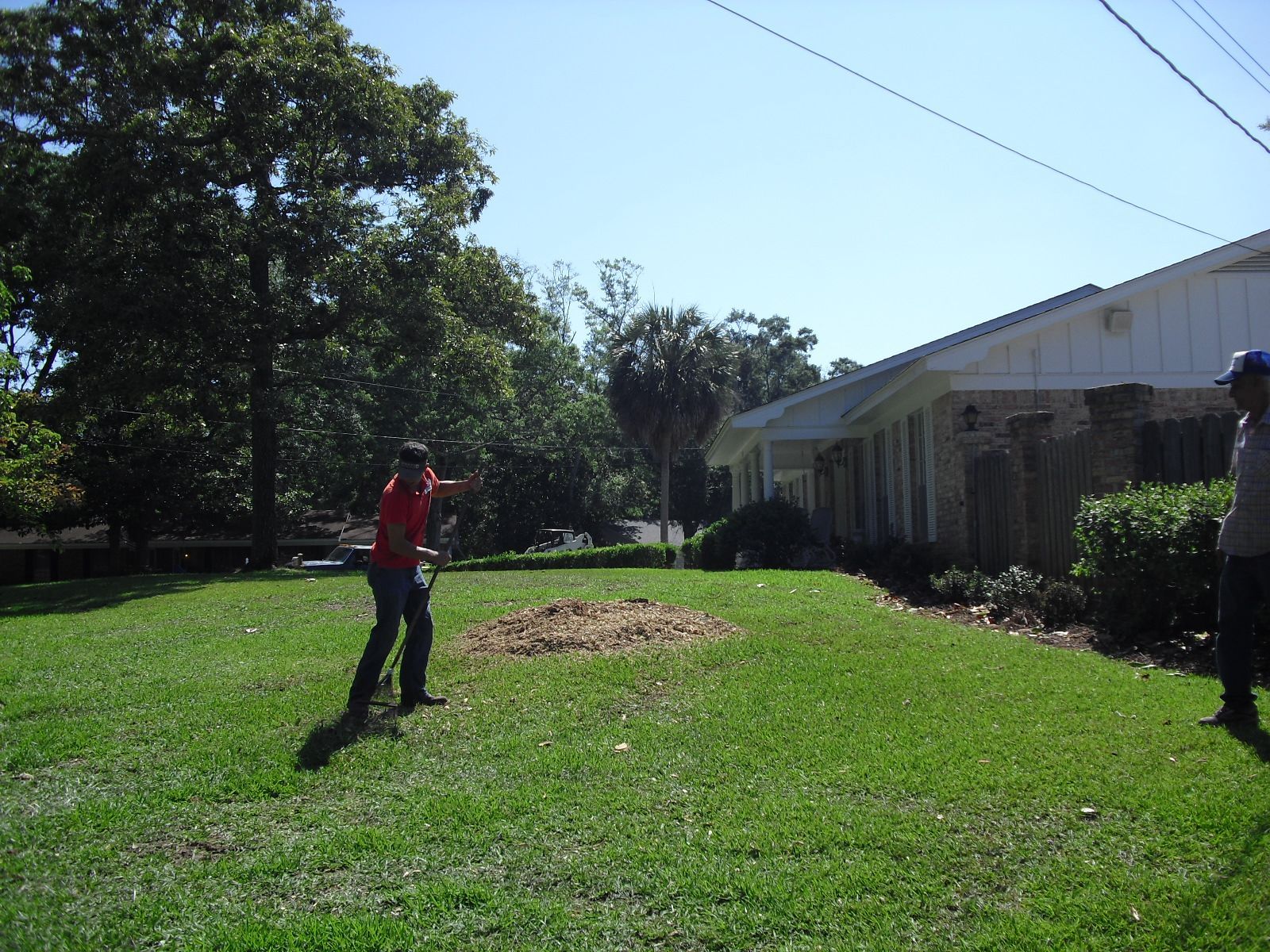 Two people stand on a grassy lawn near a house, with one gesturing toward a pile of mulch or debris in the yard.