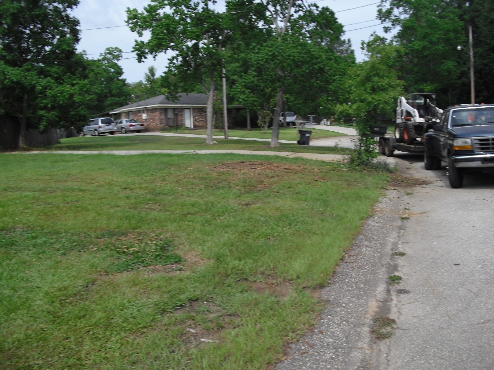 A yard with a patch of dirt sits near a road with a parked truck towing a construction vehicle near a residential house.