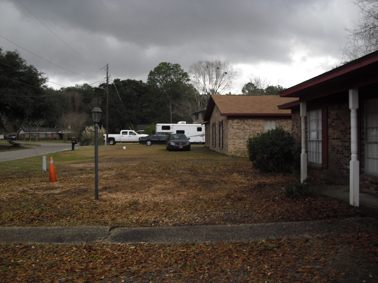 A view of a yard with scattered leaves, several parked vehicles, a camper, and a brick house under a cloudy sky.