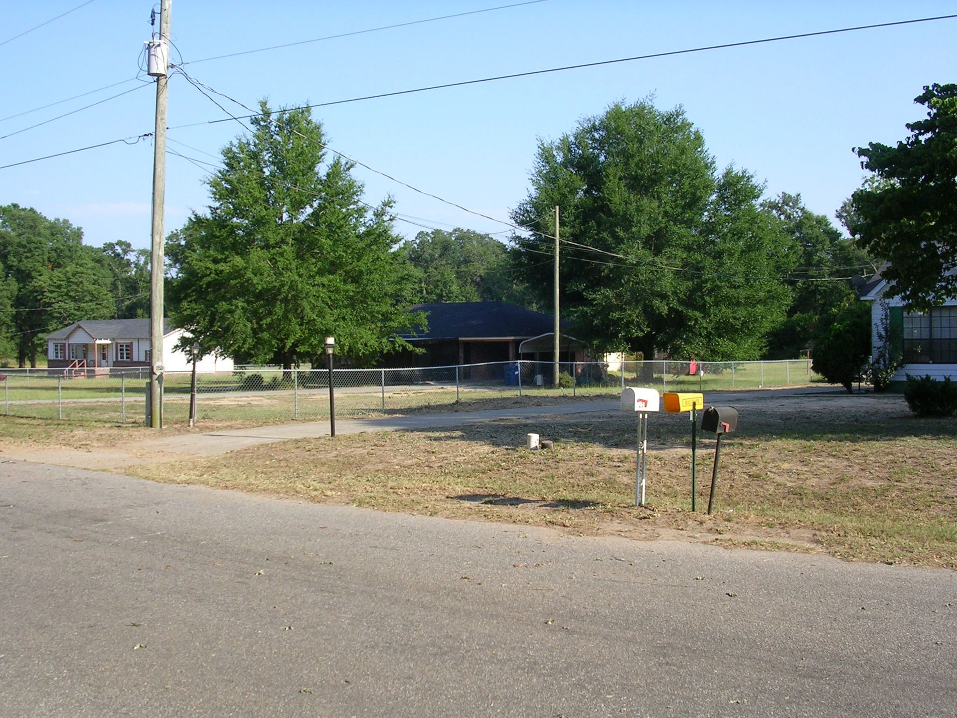 Rural street scene featuring houses, green trees, a utility pole, and a row of mailboxes along a gravel driveway.