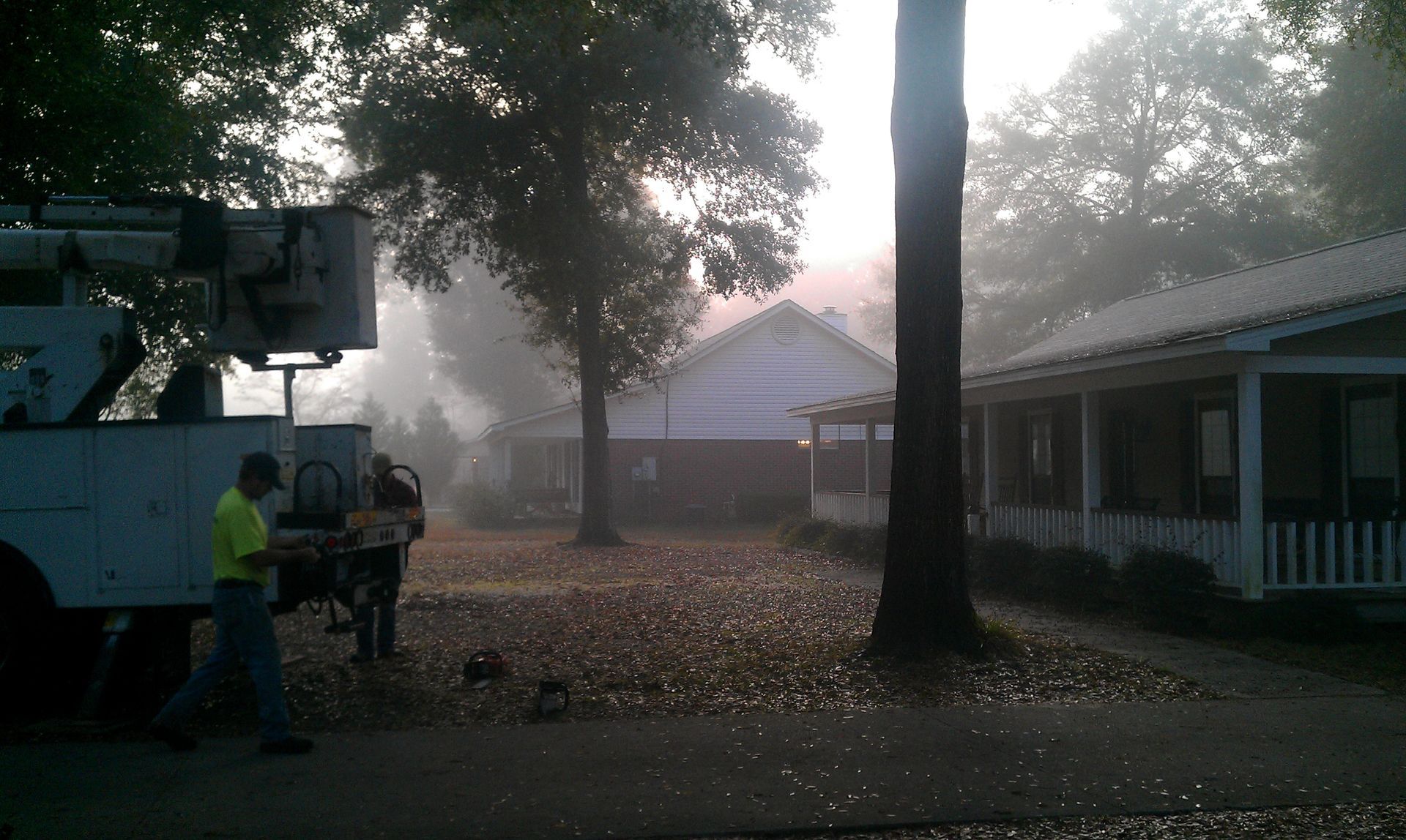 A worker in a yellow shirt stands near a utility truck parked in a foggy, leaf-covered yard between two houses.