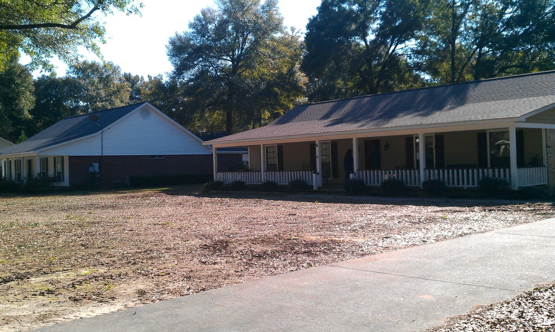A single-story brick house with a wide front porch and an attached garage under a canopy of green trees.