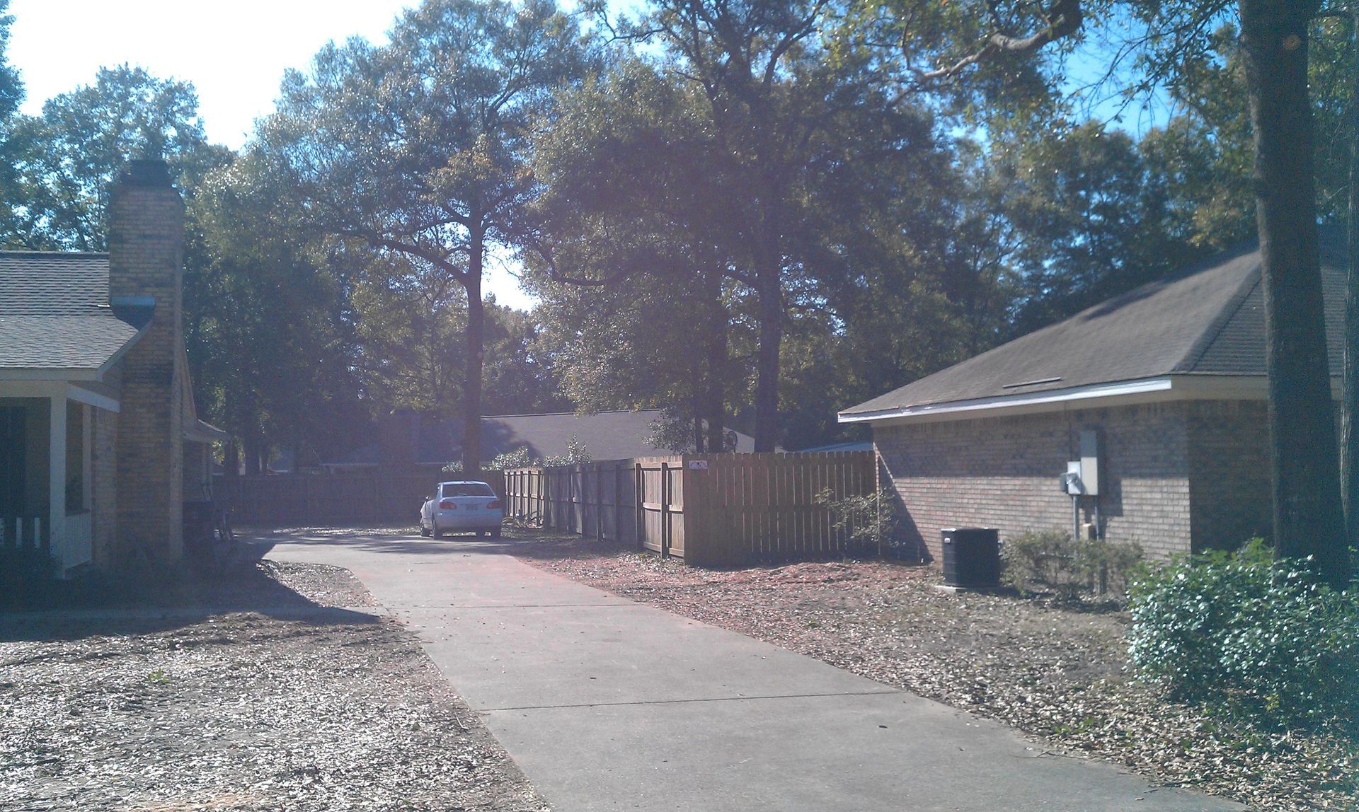 A paved driveway leads between two brick houses toward a wooden fence and a parked white car under large, leafy trees.