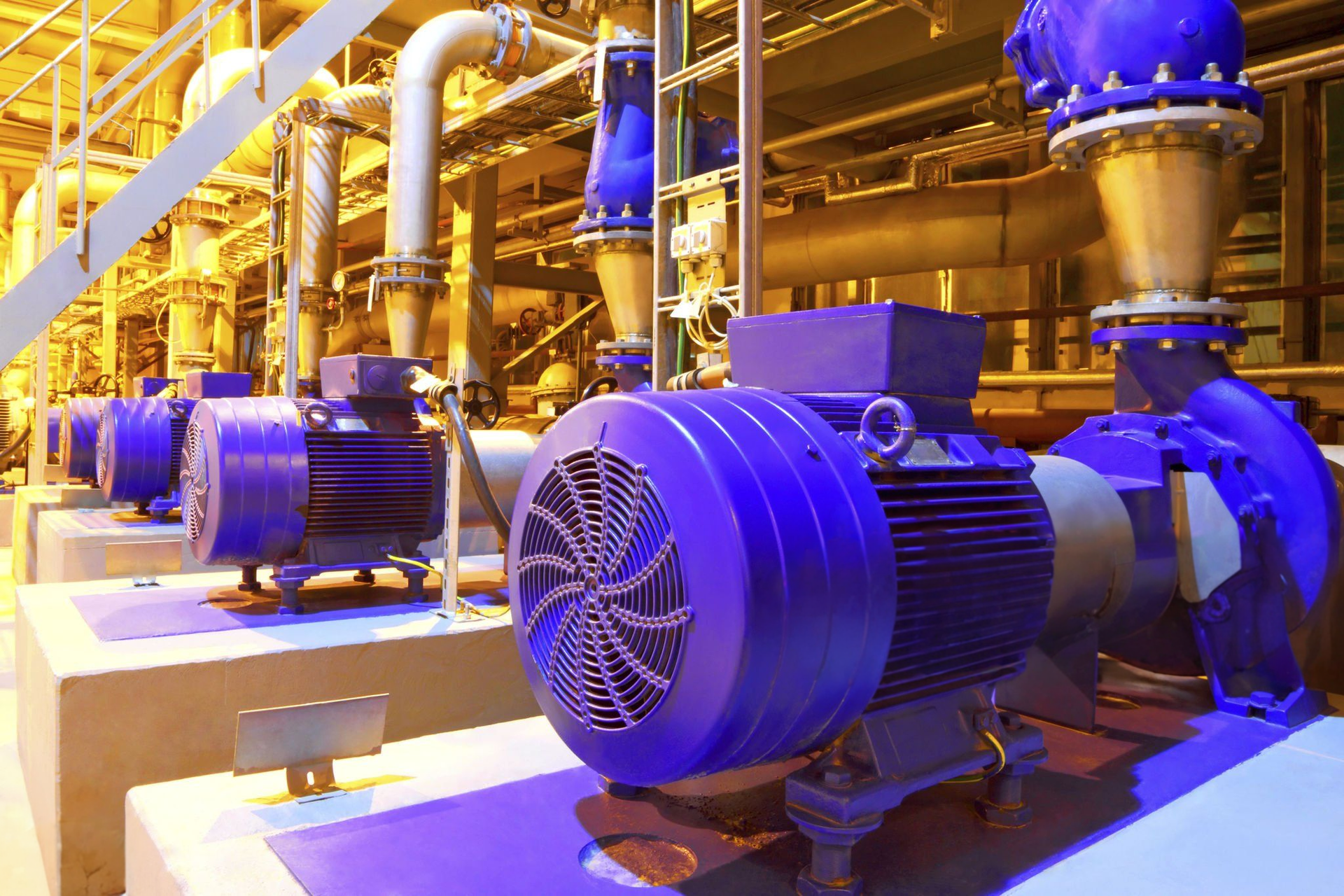 Blue industrial pumps and motors in a brightly lit factory with yellow piping and metal equipment.