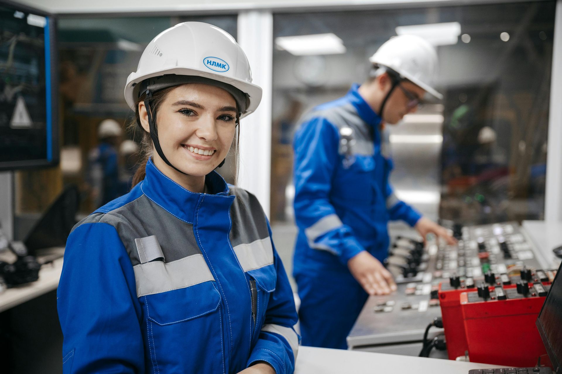 Worker in blue uniform and white hard hat smiles in a factory beside another worker and machinery