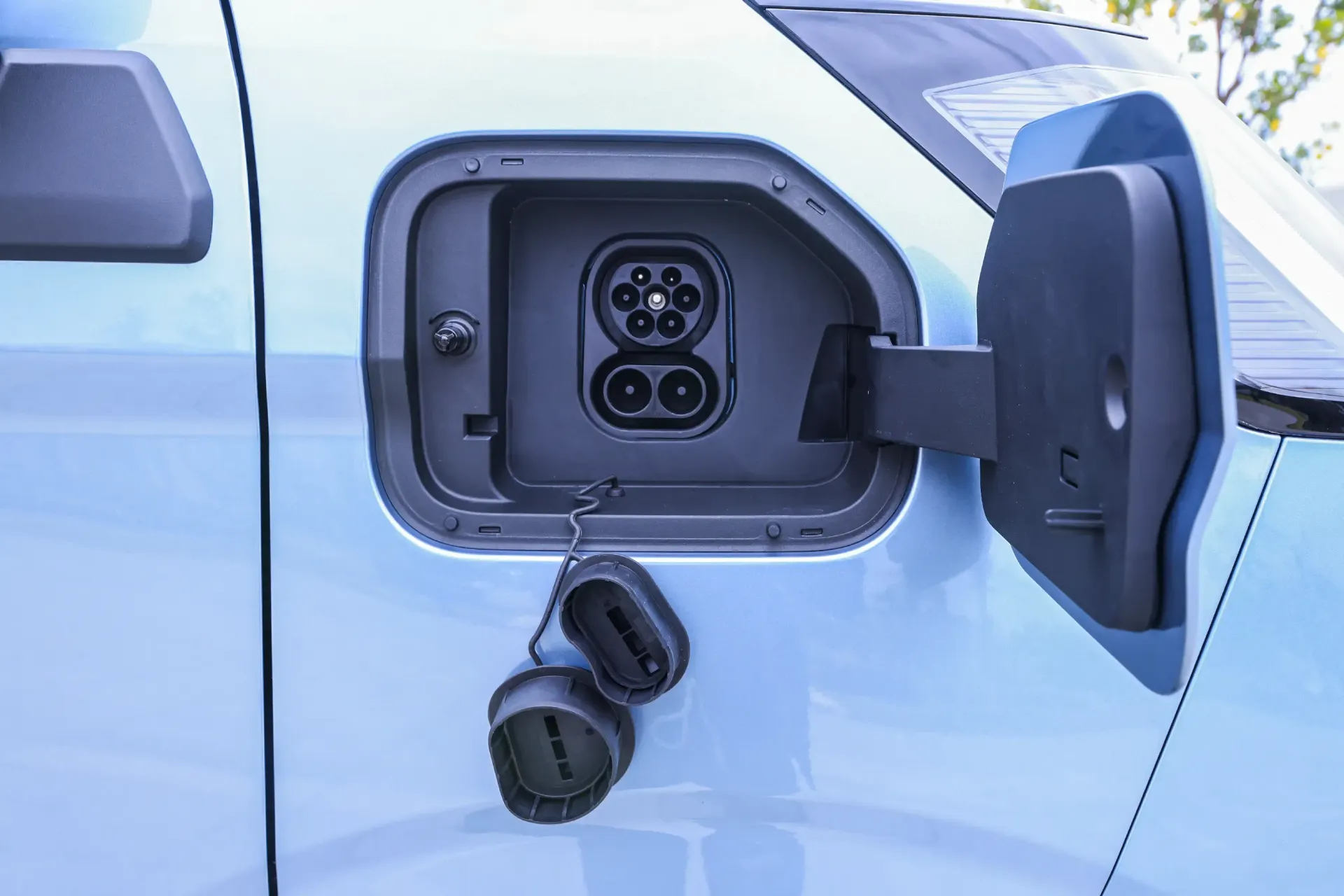Open electric vehicle charging port on the side of a light blue car, with two rubber protective caps hanging below.