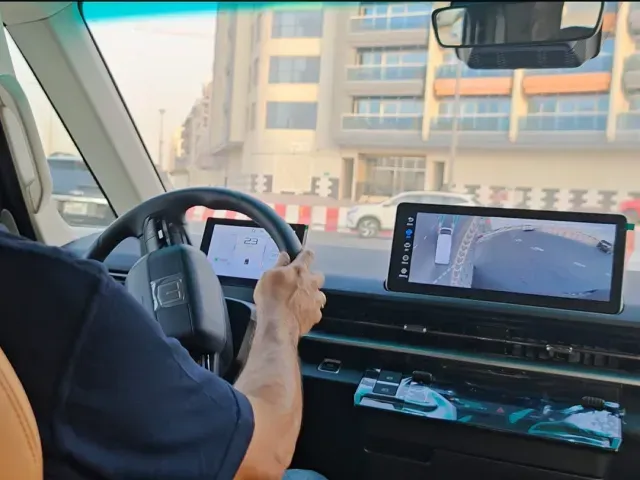 A driver's perspective from inside a vehicle, showing the steering wheel and a dashboard screen displaying a camera feed.