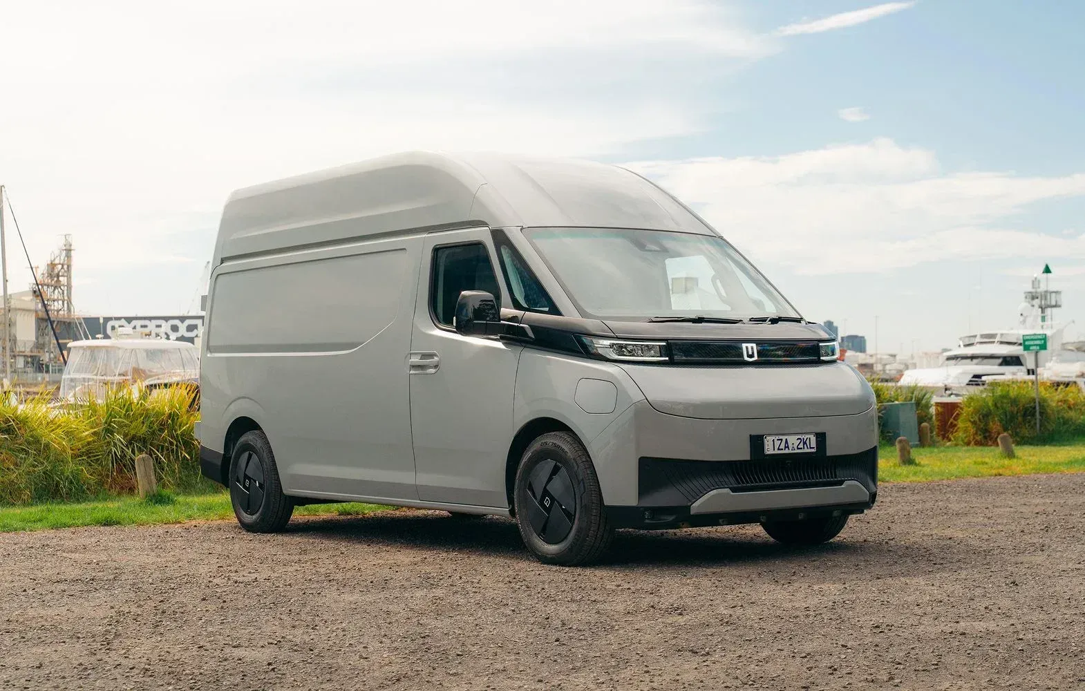 A light grey electric cargo van parked on a gravel surface outdoors with a partially cloudy sky in the background.