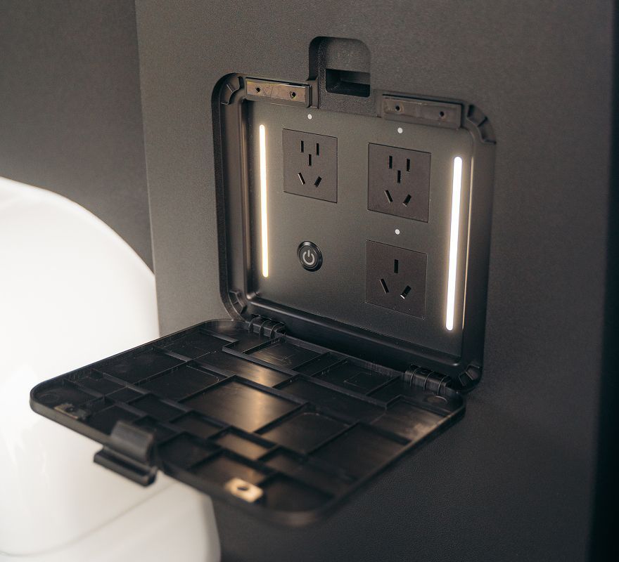 A black, wall-mounted utility panel with three power outlets and a central power button, illuminated by vertical LEDs.