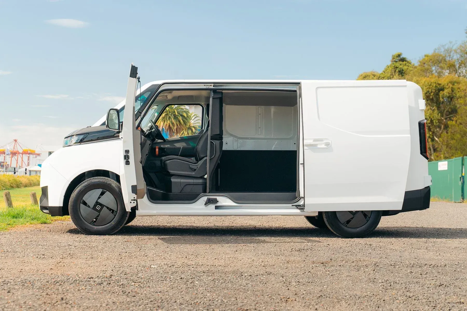 A side profile of a white electric cargo van parked on a gravel lot with its passenger door and side door open.