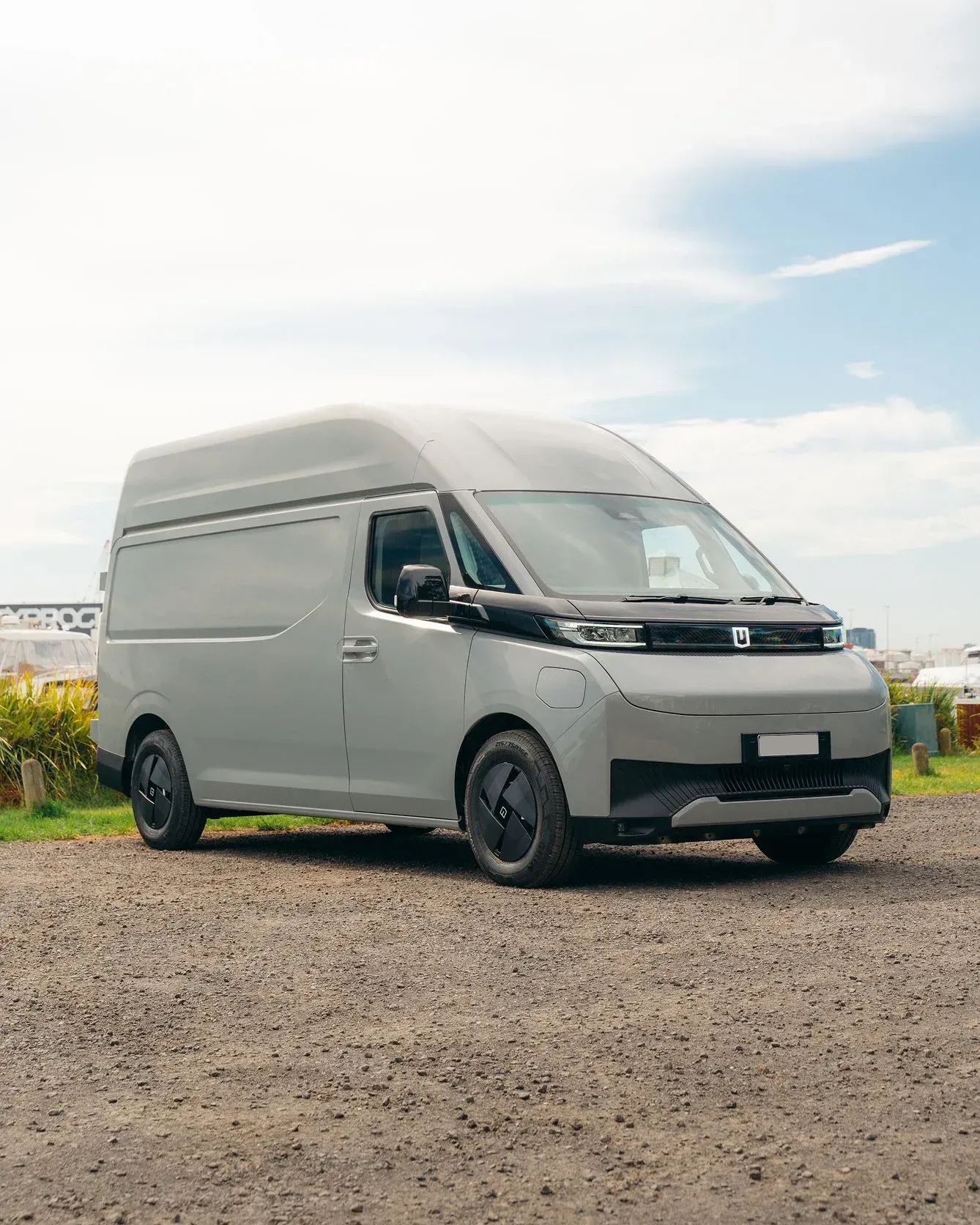 A light gray electric cargo van parked on a gravel surface under a bright sky.