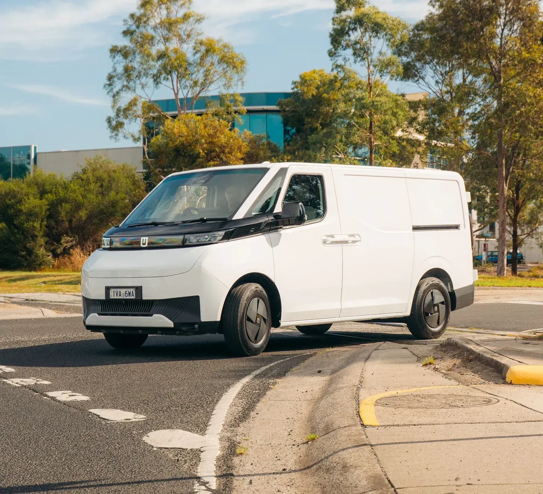 A white commercial delivery van parked on an asphalt road near trees and a modern office building on a sunny day.