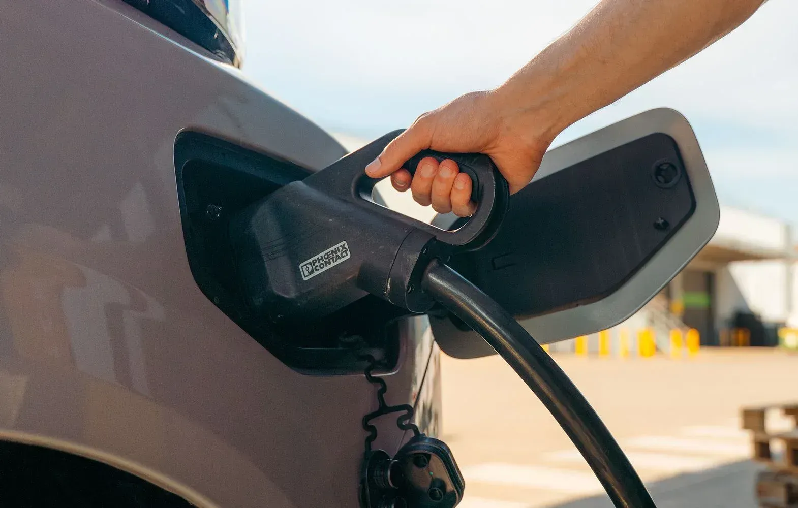 A hand plugging a charging cable into an electric vehicle's charging port outdoors.