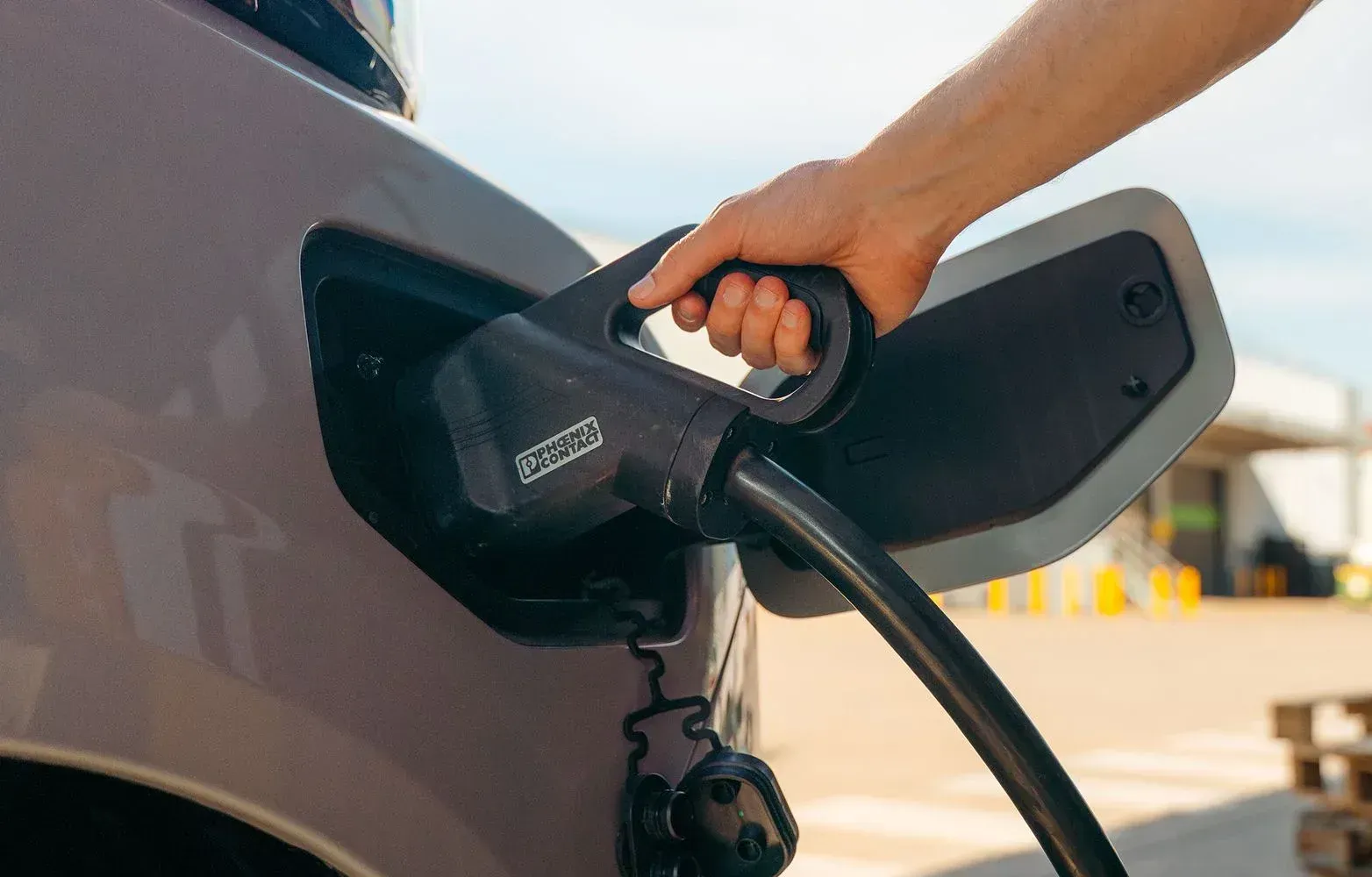 A person plugging a black electric vehicle charging cable into the side port of a light-colored car outdoors.