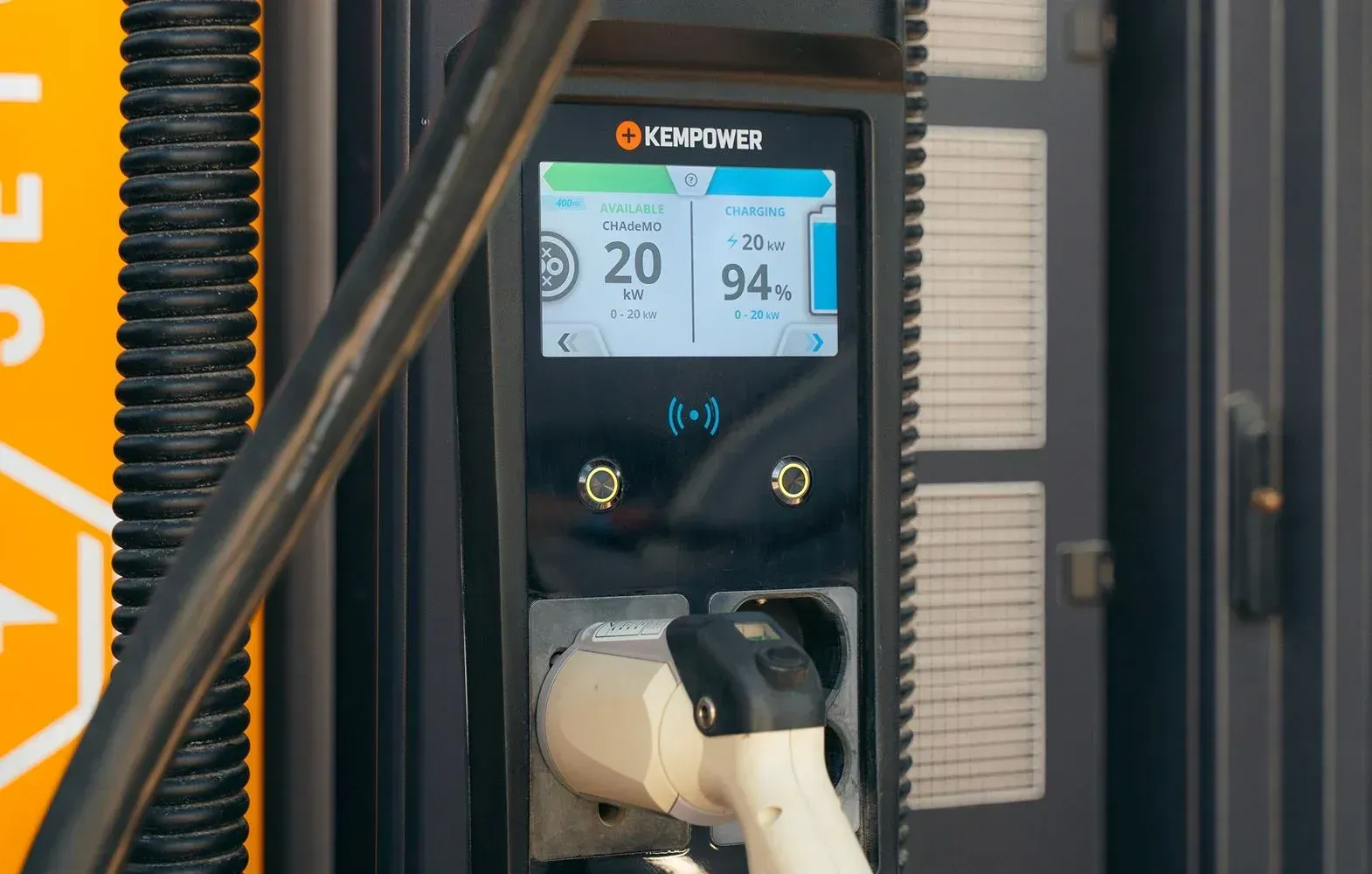 Electric vehicle charging station with a screen showing 20 kW power and a 94% charge level, with a charging cable plugged in.