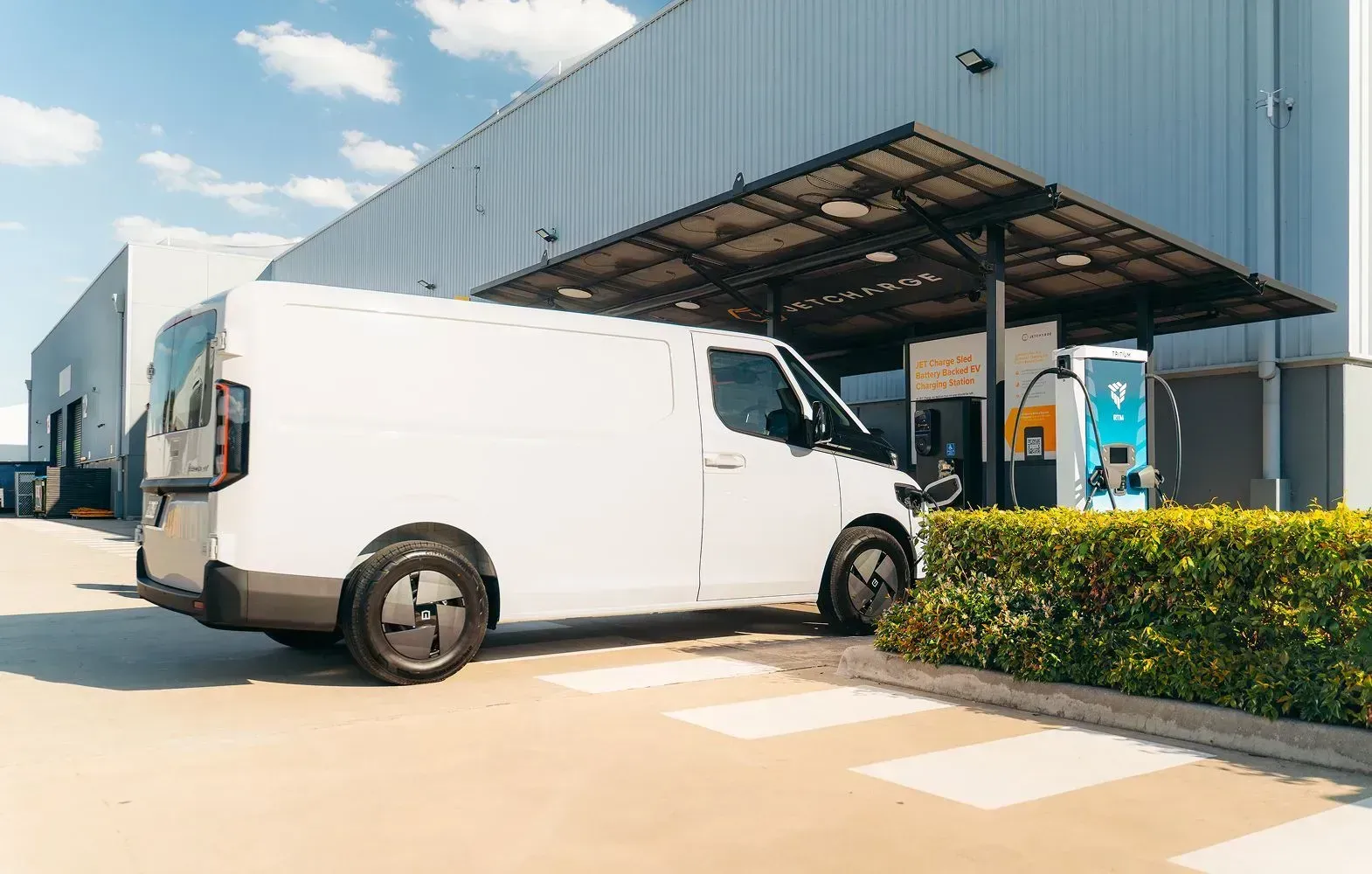 A white delivery van parked at an outdoor charging station next to a large industrial building on a sunny day.