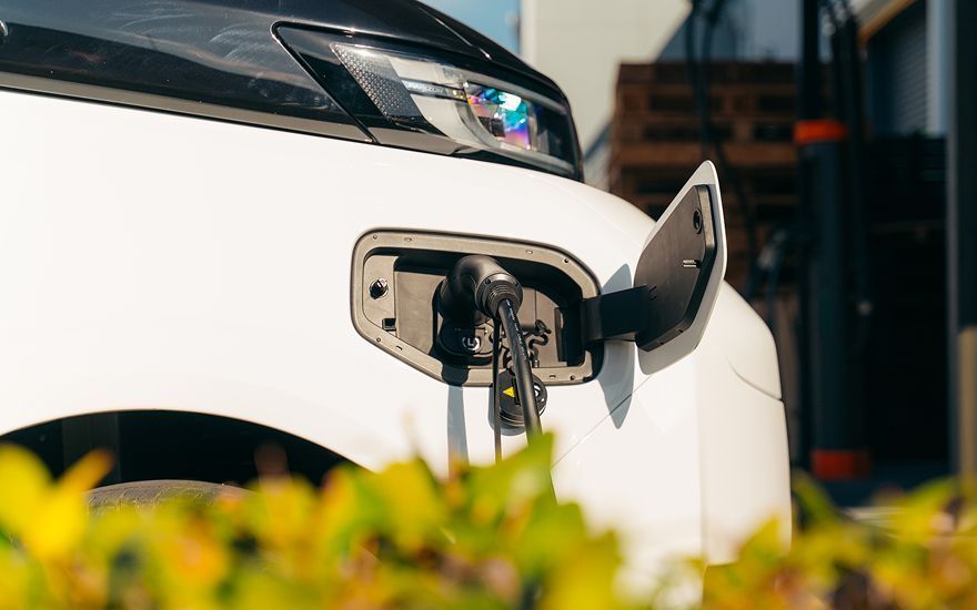 A charging cable plugged into the side of a white electric vehicle in front of a building with blurred green foliage.