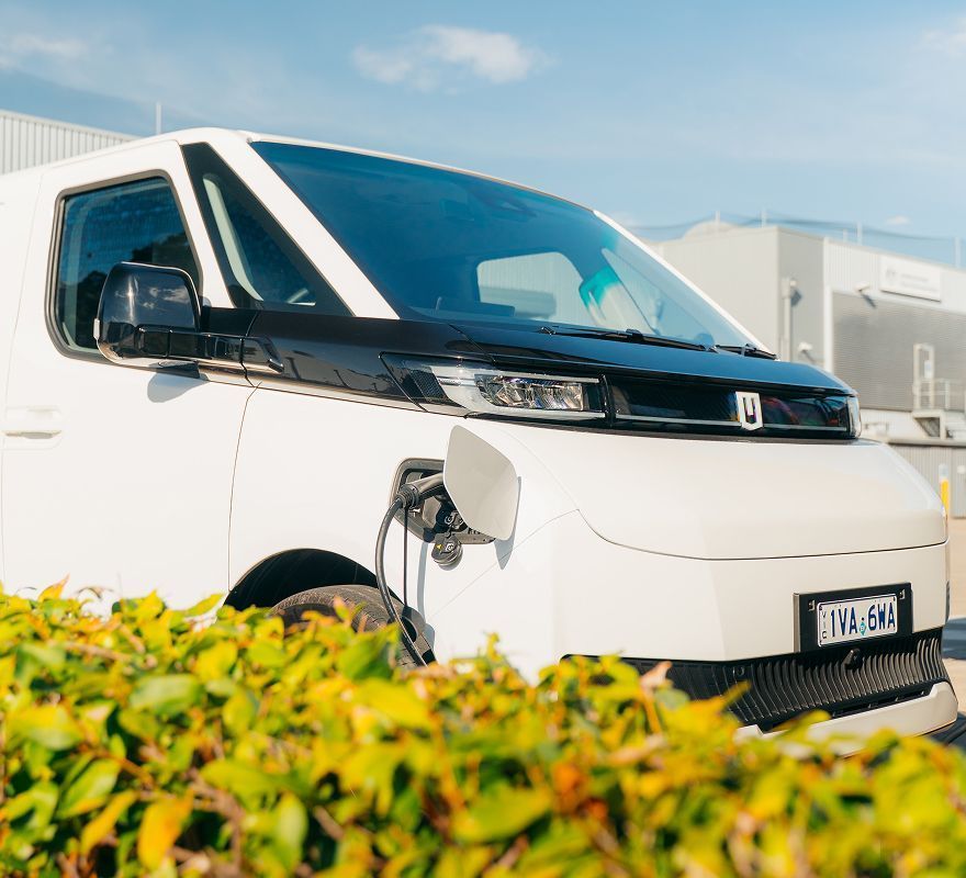 A white electric delivery van is parked outdoors and plugged into a charging station, partially obscured by green bushes.