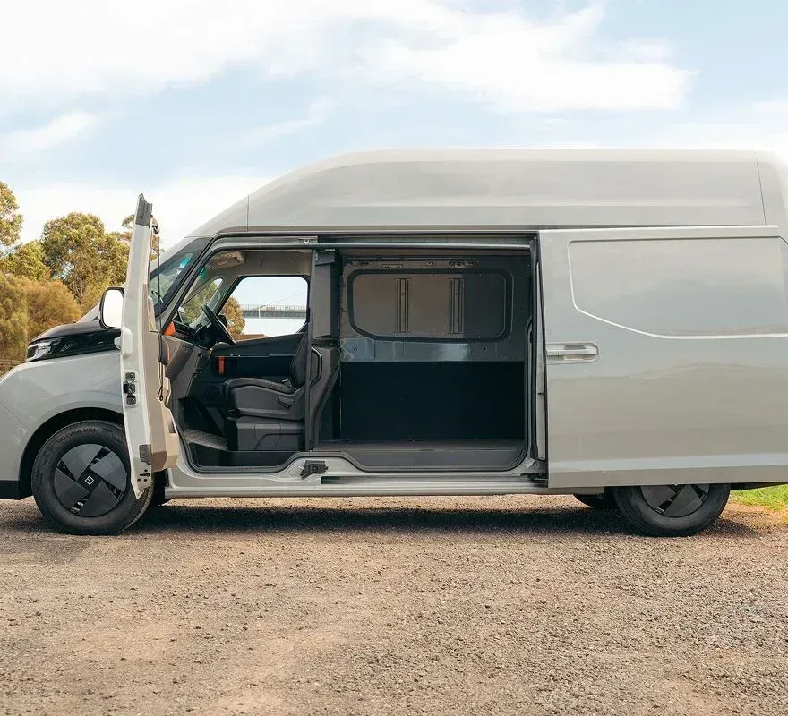 A light gray cargo van parked on a gravel surface with its side door open, revealing the driver's seat and empty interior.