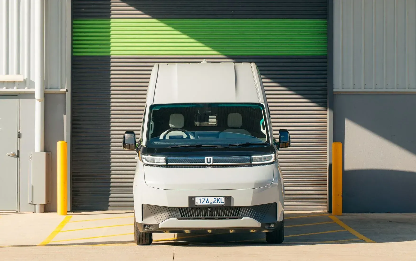 A light grey electric delivery van parked in front of a warehouse door with green and grey horizontal stripes.