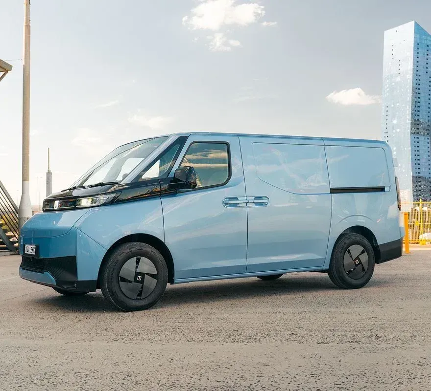 A light blue electric cargo van parked on a paved lot with a modern skyscraper in the background.