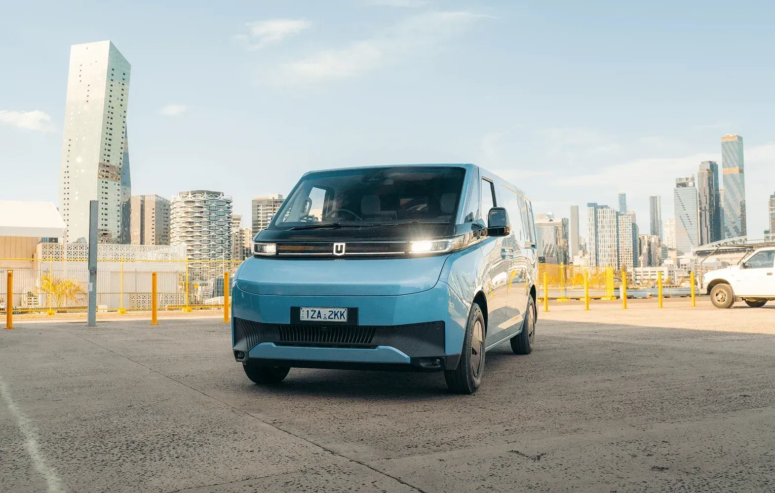 A light blue electric van is parked in a paved urban lot with city skyscrapers in the background under a sunny sky.