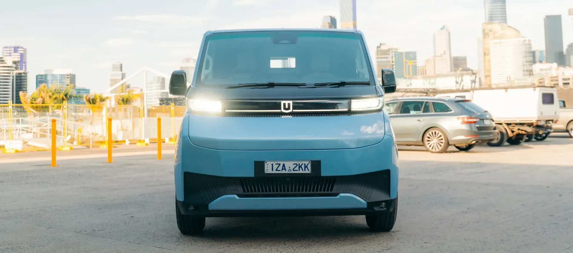 A light blue electric delivery van faces forward in an outdoor parking lot with a city skyline in the background.