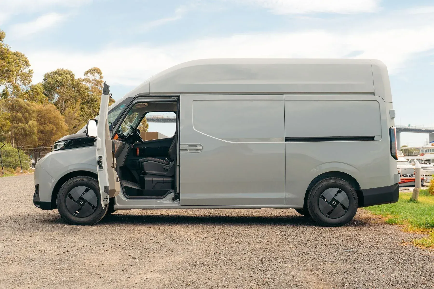 A gray cargo van parked on a gravel path with its driver-side door open, seen from the side.