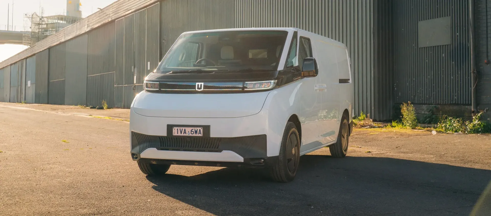 A white electric commercial van parked on an asphalt lot beside a corrugated metal building at sunrise.