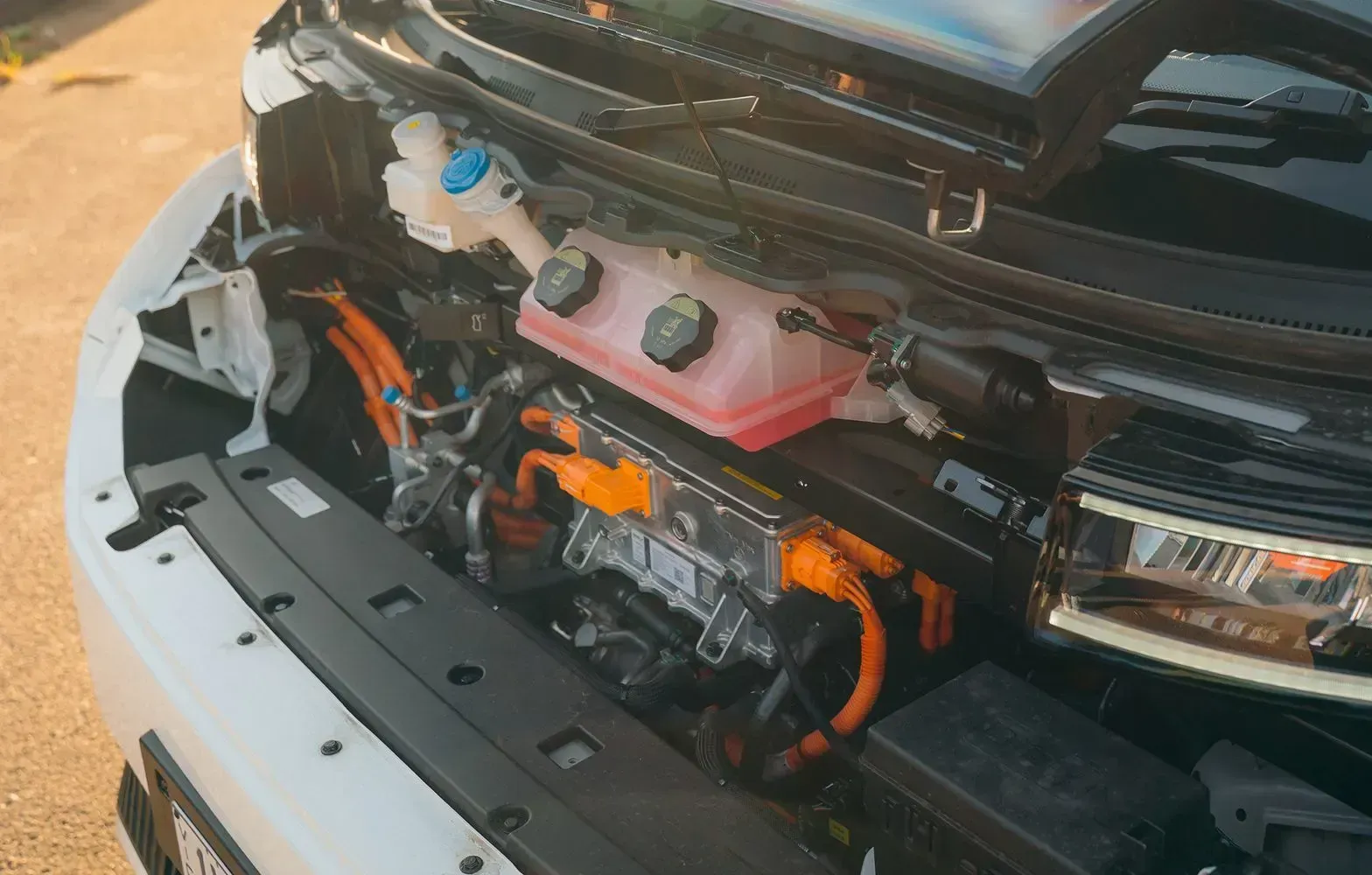 An open engine bay of an electric vehicle showing a translucent coolant reservoir and bright orange high-voltage cables.
