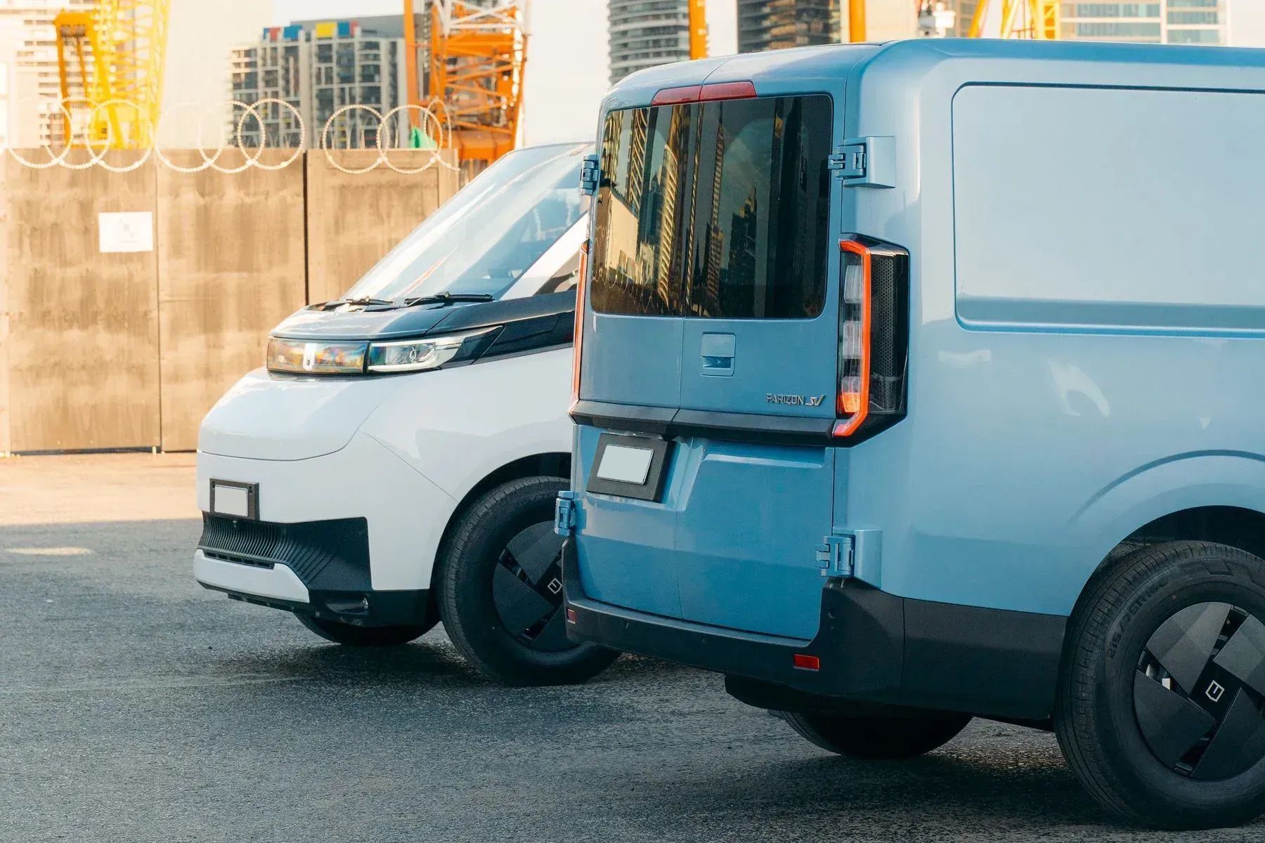 A white and a blue electric cargo van parked side-by-side in an outdoor industrial lot.