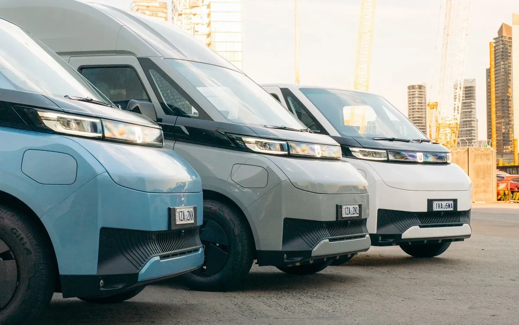 A row of three electric vans in light blue, gray, and white parked in a city lot with tall buildings in the background.
